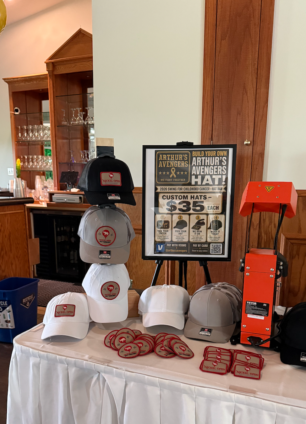 Custom Hat Bar table setup with hats, patches, a heat press, and event signage